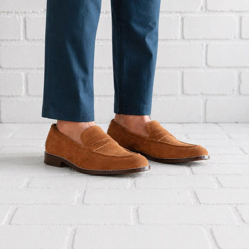 Op de afbeelding: Een paar bruine su&egrave;de penny loafers. De schoenen hebben een klassiek ontwerp met een ronde neus en een leren zool. De loafers zijn warmbruin van kleur en lijken ongedragen.