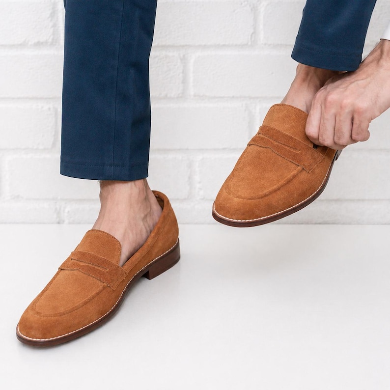 Op de afbeelding: Bruine penny loafers van su&egrave;de met een leren zool. De schoenen hebben een klassiek ontwerp met een band over de bovenkant en worden getoond met een marineblauwe broek.