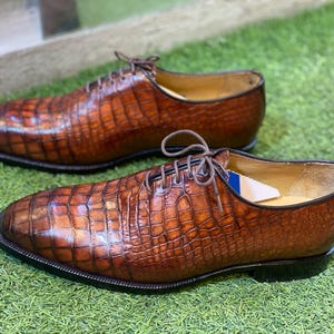 May include: A pair of brown leather dress shoes with a crocodile skin texture. The shoes have a classic lace-up design, a black sole, and a light-colored interior. The shoes are displayed on a green surface.