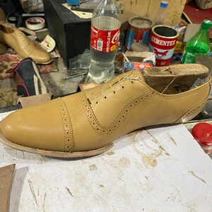 K&ouml;nnte beinhalten: Ein hellbrauner Lederschuh im Herstellungsprozess, mit einem h&ouml;lzernen Schuhleisten im Inneren. Der Schuh hat dekorative Perforationen und ein klassisches Cap-Toe-Design. Der Schuh liegt auf einer wei&szlig;en Oberfl&auml;che.