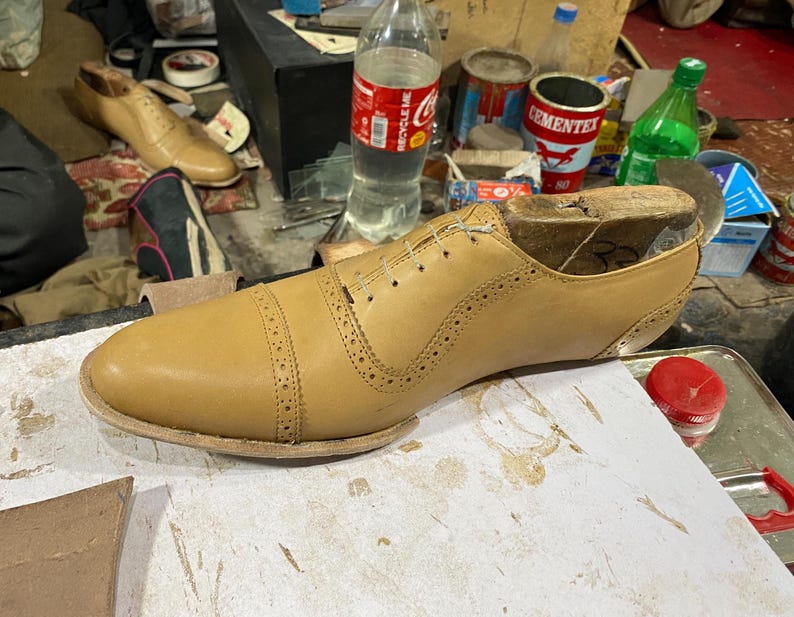 K&ouml;nnte beinhalten: Ein hellbrauner Lederschuh im Herstellungsprozess, mit einem h&ouml;lzernen Schuhleisten im Inneren. Der Schuh hat dekorative Perforationen und ein klassisches Cap-Toe-Design. Der Schuh liegt auf einer wei&szlig;en Oberfl&auml;che.