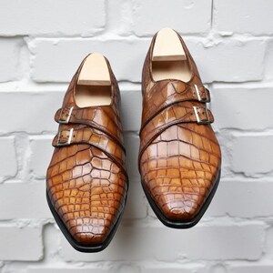 May include: A pair of brown leather dress shoes with a crocodile skin texture. The shoes have two straps with silver buckles and a pointed toe. The shoes are displayed on a white brick background.