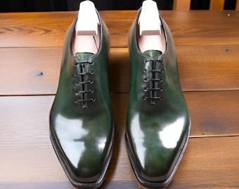Puede incluir: Un par de zapatos de vestir de cuero verde con cordones negros y un acabado pulido. Los zapatos tienen un diseño Oxford clásico con punta afilada y se exhiben con hormas de madera.
