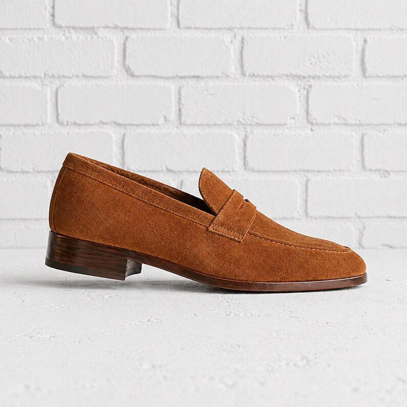 Op de afbeelding: Een bruine su&egrave;de loafer met een lage hak. De schoen heeft een klassiek ontwerp met een penny band over de wreef. De zool en hak zijn donkerder bruin. De schoen staat tegen een witte bakstenen muur.