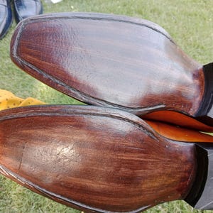 Puede incluir: Primer plano de las suelas de dos zapatos de vestir de cuero marr&oacute;n. Las suelas son de un marr&oacute;n oscuro intenso con vetas de madera visibles y un tac&oacute;n negro. Los zapatos son probablemente nuevos o bien cuidados.