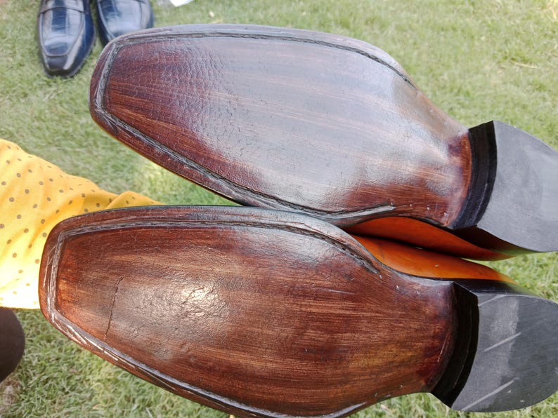Puede incluir: Primer plano de las suelas de dos zapatos de vestir de cuero marr&oacute;n. Las suelas son de un marr&oacute;n oscuro intenso con vetas de madera visibles y un tac&oacute;n negro. Los zapatos son probablemente nuevos o bien cuidados.