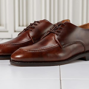 Peut inclure: Une paire de chaussures de ville en cuir marron avec une finition brillante. Les chaussures ont un design classique avec un bout arrondi, une fermeture à lacets et un petit talon. Les chaussures sont présentées sur une surface carrelée blanche.