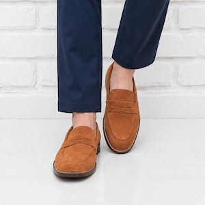 Op de afbeelding: Een paar bruine penny loafers van su&egrave;de. De schoenen hebben een klassiek ontwerp met een ronde neus, een band over de wreef en een gestikte zool. De loafers worden gedragen met een marineblauwe broek.