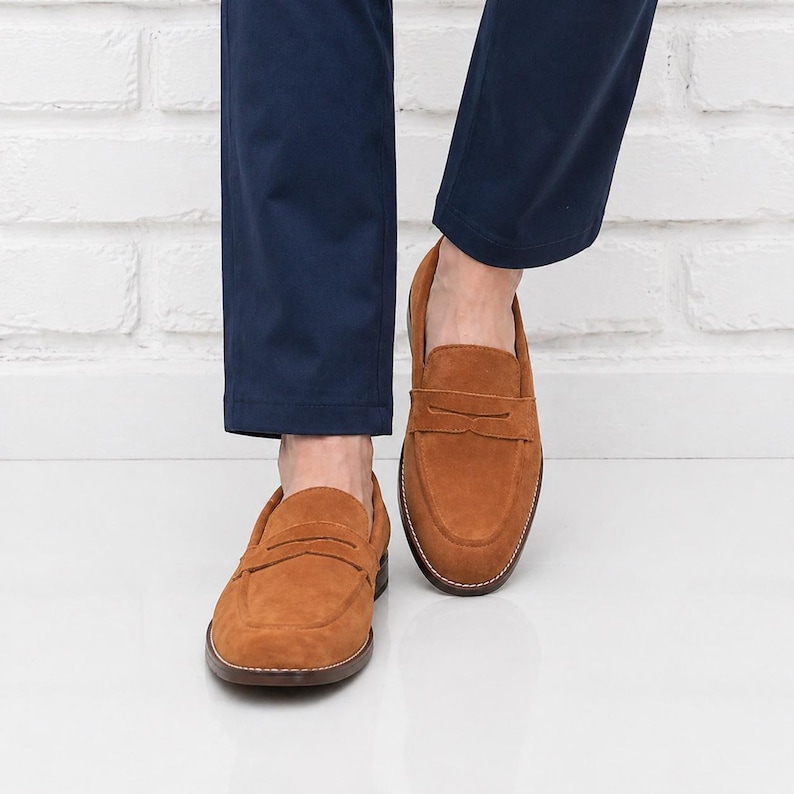 Op de afbeelding: Een paar bruine penny loafers van su&egrave;de. De schoenen hebben een klassiek ontwerp met een ronde neus, een band over de wreef en een gestikte zool. De loafers worden gedragen met een marineblauwe broek.