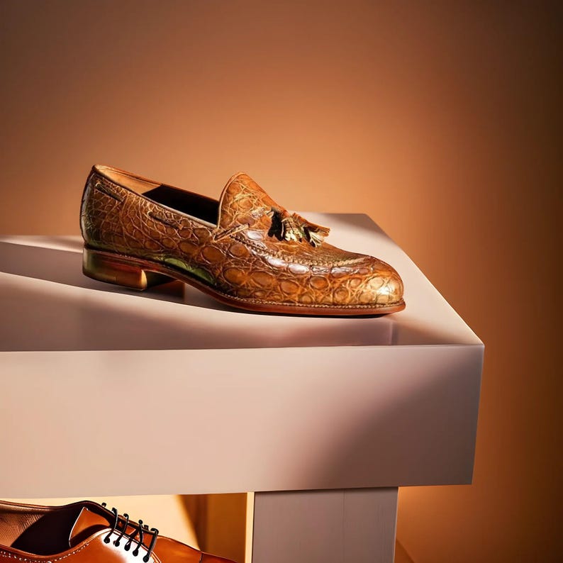 May include: A brown leather loafer with a crocodile texture, featuring a tassel detail and a low heel. The shoe is displayed on a white surface against a warm, neutral background, showcasing its polished finish.