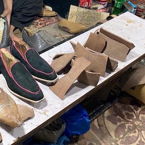 K&ouml;nnte beinhalten: Werkstatt eines Schuhmachers mit verschiedenen Lederschuhkomponenten und Werkzeugen. Mehrere Schuhpaare befinden sich in verschiedenen Stadien der Fertigstellung, darunter schwarze Wildleder-Loafer mit rosa Besatz. Holzschuhleisten und Lederausschnitte sind ebenfalls sichtbar.