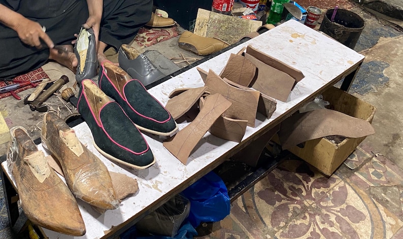 K&ouml;nnte beinhalten: Werkstatt eines Schuhmachers mit verschiedenen Lederschuhkomponenten und Werkzeugen. Mehrere Schuhpaare befinden sich in verschiedenen Stadien der Fertigstellung, darunter schwarze Wildleder-Loafer mit rosa Besatz. Holzschuhleisten und Lederausschnitte sind ebenfalls sichtbar.