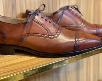 May include: A pair of brown leather Oxford shoes with decorative perforations and dark brown laces. The shoes are displayed on a wooden shelf with a gold trim. The shoes have a classic, formal design.