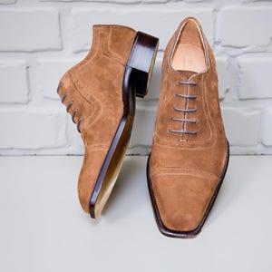 May include: A pair of brown suede dress shoes with a square toe and dark brown soles. The shoes have a lace-up design and a stacked heel. The background is a white brick wall.
