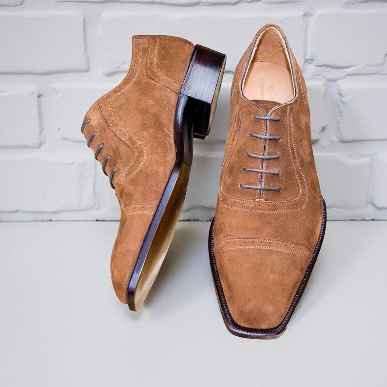 May include: A pair of brown suede dress shoes with a square toe and dark brown soles. The shoes have a lace-up design and a stacked heel. The background is a white brick wall.