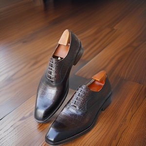K&ouml;nnte beinhalten: Ein Paar braune Lederschuhe mit Krokodilleder-Textur. Die Schuhe haben ein klassisches Schn&uuml;rdesign und werden auf einer Holzoberfl&auml;che pr&auml;sentiert. In jedem Schuh sind Schuhspanner eingesetzt.