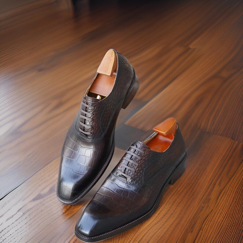 K&ouml;nnte beinhalten: Ein Paar braune Lederschuhe mit Krokodilleder-Textur. Die Schuhe haben ein klassisches Schn&uuml;rdesign und werden auf einer Holzoberfl&auml;che pr&auml;sentiert. In jedem Schuh sind Schuhspanner eingesetzt.
