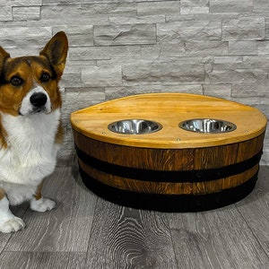 May include: A wooden dog bowl stand made from a repurposed barrel with two stainless steel bowls. A corgi dog is sitting in front of the stand.