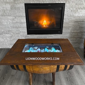 Peut inclure: Une table basse en bois de baril avec un plateau en verre qui révèle un refroidisseur intégré pour le stockage de bouteilles. La table a une finition brun foncé et des bandes métalliques noires autour du baril. Le texte "LIONWOODWORKS.COM" est imprimé sur la surface de la table.
