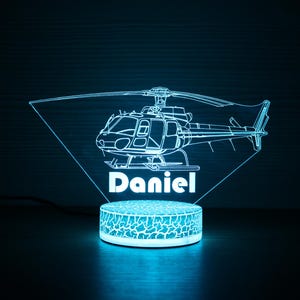 May include: A blue and white 3D printed night light shaped like a helicopter with the name "Daniel" underneath. The light is on a round base with a textured surface.