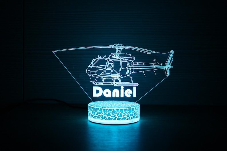 May include: A blue and white 3D printed night light shaped like a helicopter with the name "Daniel" underneath. The light is on a round base with a textured surface.