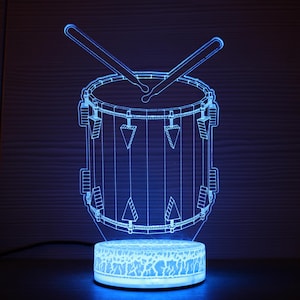 以下が含まれることがあります： ドラムスティック付きの青いLEDドラムランプ。ランプは、丸いベースを備えたドラムの詳細なアウトラインを備えています。ドラムスティックはドラムの上に配置されています。ランプは明るい青色の光で照らされています。