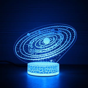 Puede incluir: Una luz nocturna azul con temática de galaxia con una base blanca. La luz presenta un diseño detallado de una galaxia con planetas y estrellas.