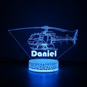 May include: A blue 3D printed night light shaped like a helicopter with the name "Daniel" underneath. The light is on a white base with a textured surface.
