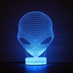 May include: A blue 3D printed alien head night light with a white base. The light is on and illuminating the alien's face.