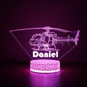 May include: A pink LED night light shaped like a helicopter with the name "Daniel" underneath. The light is on a white base with a textured surface.