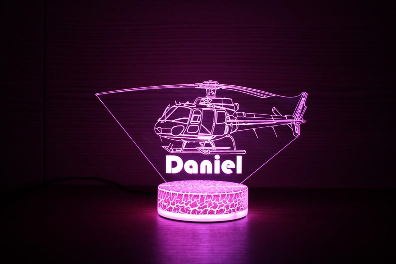 May include: A pink LED night light shaped like a helicopter with the name "Daniel" underneath. The light is on a white base with a textured surface.