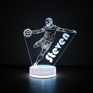 以下が含まれることがあります： サッカーボールを蹴るサッカー選手を描いた、光るLEDランプ。 選手の画像は白いアウトラインで、名前「Steven」が白い文字で表示されています。 ランプはテクスチャ加工されたベースと三角形のデザインです。