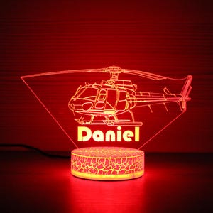 May include: A red LED night light shaped like a helicopter with the name "Daniel" underneath. The light is on a white base with a textured surface.