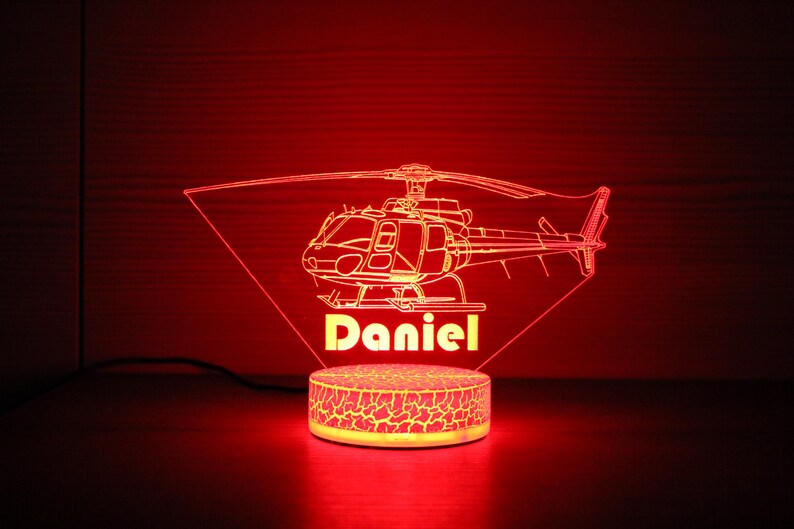 May include: A red LED night light shaped like a helicopter with the name "Daniel" underneath. The light is on a white base with a textured surface.