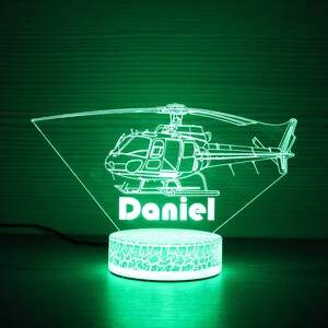May include: A green LED night light shaped like a helicopter with the name "Daniel" underneath. The light is on a white base with a textured surface.
