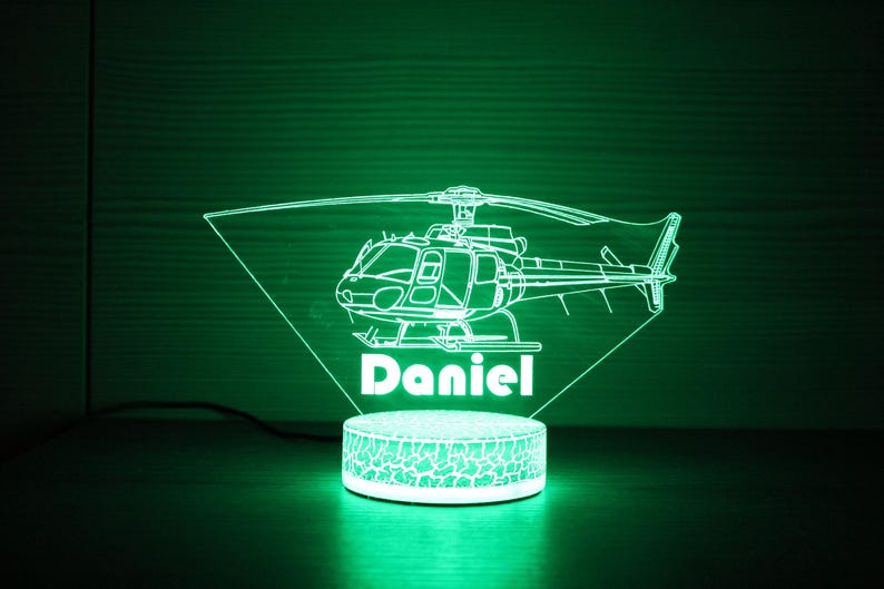 May include: A green LED night light shaped like a helicopter with the name "Daniel" underneath. The light is on a white base with a textured surface.