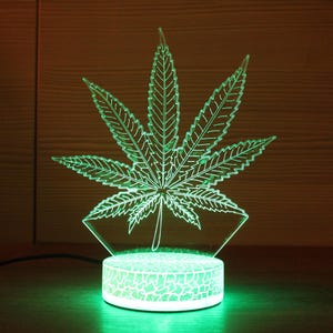 May include: A green LED night light shaped like a marijuana leaf. The light is on and illuminating the leaf.