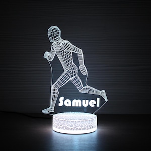 Puede incluir: Una luz nocturna blanca impresa en 3D con forma de persona corriendo con el nombre "Samuel" impreso debajo. La luz está sobre una base blanca con una superficie texturizada.