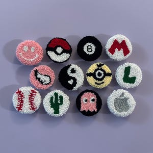 May include: A collection of twelve round, textured patches with various designs. The patches include a smiley face, a Pokeball, an eight ball, the letter "M", a yin-yang symbol, a Minion, the letter "L", a baseball, a cactus, a ghost, and an apple.