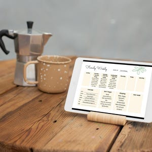 Könnte beinhalten: Ein digitales Tablet mit einem wöchentlichen Familienplaner, das auf einem Holzständer steht. Eine silberne Espressokanne und eine gesprenkelte braune Tasse stehen auf einem Holztisch. Der Bildschirm des Tablets zeigt einen Zeitplan.