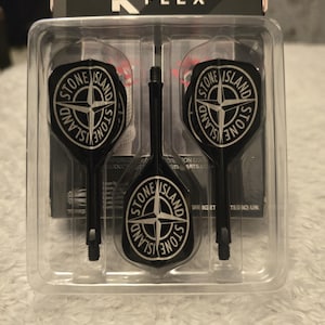May include: A set of three black dart flights with the Stone Island logo in a clear plastic package. The logo is a compass rose design in white, set against a black background. The package is labeled "K FLEX".