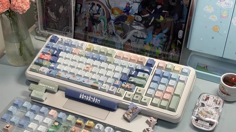 May include: A white mechanical keyboard with pastel blue, green, and pink keycaps featuring various illustrations. The keyboard has a blue wrist rest with the word "TokkiTo" printed on it. The keyboard is surrounded by other keycaps and a small clear container with a decorative design.