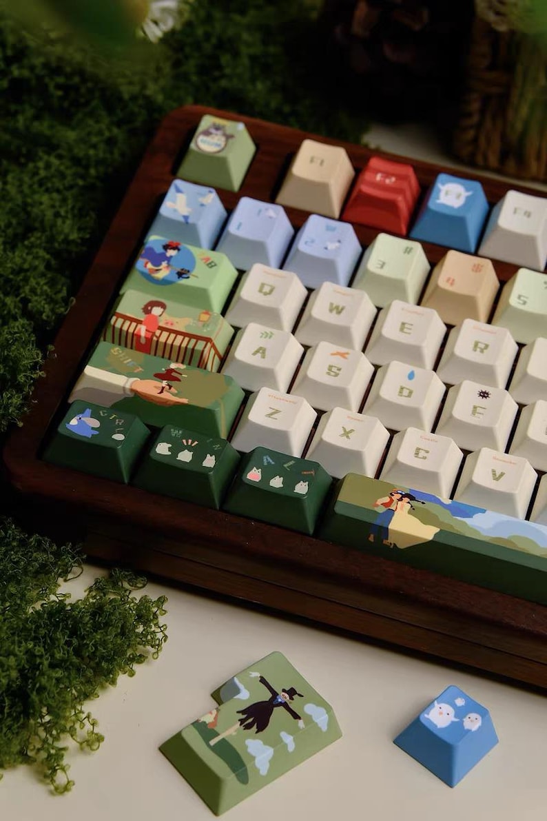 K&ouml;nnte beinhalten: Eine benutzerdefinierte Tastatur mit gr&uuml;nen, blauen und wei&szlig;en Tastenkappen, die Illustrationen von Figuren aus dem Animationsfilm 'Howls bewegliches Schloss' zeigen. Die Tastatur hat einen Holzrahmen und ist von gr&uuml;nem Moos umgeben.