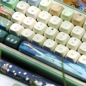 Può includere: Una tastiera verde e bianca con tasti personalizzati che presentano illustrazioni di natura e animali. La tastiera ha una barra spaziatrice con un'illustrazione blu e bianca di una barca su un'onda.