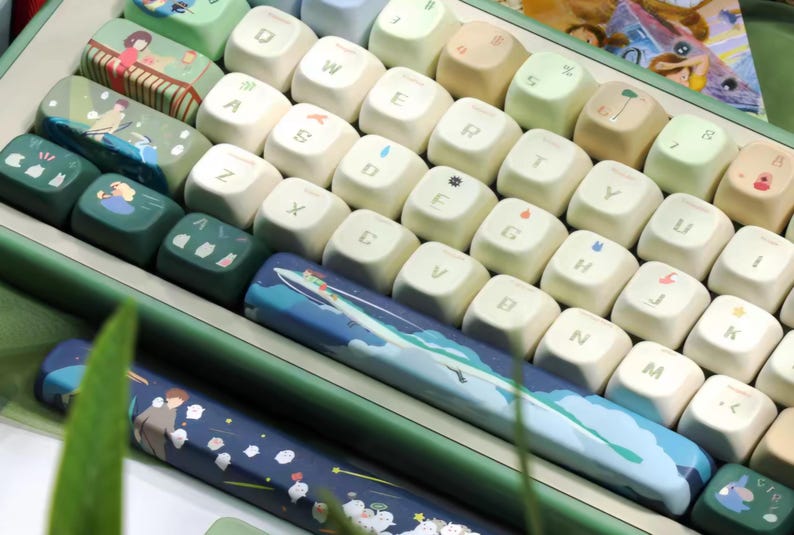 K&ouml;nnte beinhalten: Eine gr&uuml;n-wei&szlig;e Tastatur mit benutzerdefinierten Tastenkappen, die Illustrationen von Natur und Tieren zeigen. Die Tastatur hat eine Leertaste mit einer blau-wei&szlig;en Illustration eines Bootes auf einer Welle.