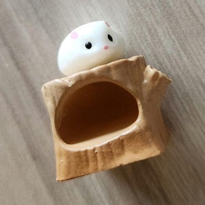 May include: A small white ceramic hamster figurine sitting on a brown ceramic tree stump with a hollowed out space.