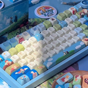 May include: A blue and white keyboard with custom keycaps featuring cartoon characters and a green and white landscape scene. The keyboard has a blue and white landscape scene on the space bar.