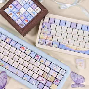 May include: Two custom keyboards with pastel blue and white keycaps featuring illustrations of anime characters. The keyboards have a light blue and white color scheme and are surrounded by decorative elements.