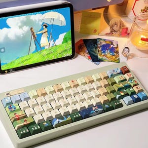 K&ouml;nnte beinhalten: Eine hellgr&uuml;ne mechanische Tastatur mit benutzerdefinierten Tastenkappen, die Illustrationen aus dem Animationsfilm 'Kikis kleiner Lieferservice' zeigen. Die Tastenkappen sind in verschiedenen Gr&uuml;nt&ouml;nen, Blau- und Beiget&ouml;nen gehalten, mit einigen wei&szlig;en Tasten. Die Tastatur hat einen hellgr&uuml;nen Rahmen und eine wei&szlig;e Basis.
