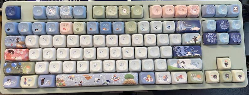 May include: A white mechanical keyboard with custom keycaps featuring illustrations from the animated film 'Howl's Moving Castle'. The keycaps are decorated with characters, scenes, and objects from the movie, including Howl, Sophie, Calcifer, and the moving castle itself.